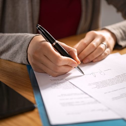 Close up of hands of young woman signing legal documents on desk. Close up of woman hand holding pen and signing legal paper seated at desk. Detail of lawyer filling official document, deal done.